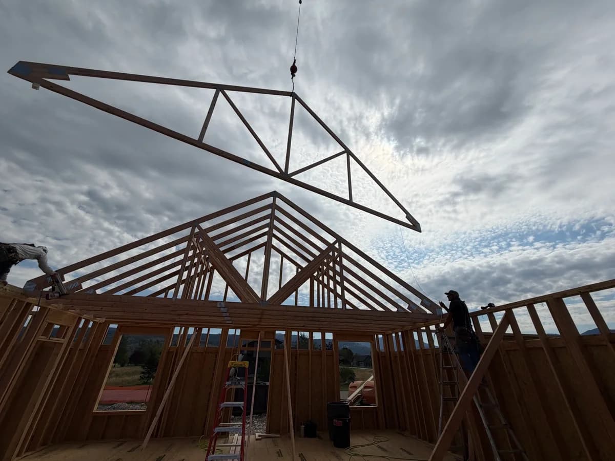 A timber roof truss being lowered onto a framed house beneath a cloudy sky